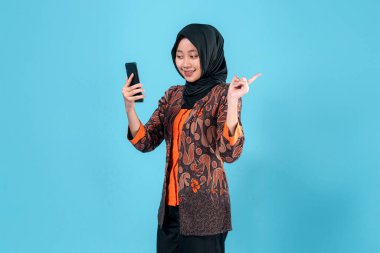Zeki bir telefon tutarken tesettürlü, gülümseyen ve yandan işaret eden kendinden emin genç Asyalı Müslüman kadın, parlak mavi bir arka plana karşı heyecan ve pozitifliğini dile getiriyor..