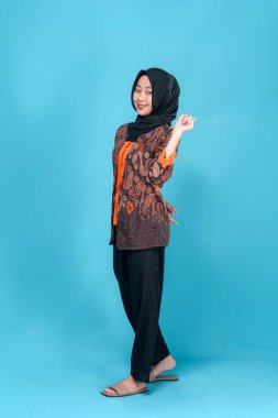 Neşeli Asyalı genç Müslüman kadın, tek parmakla yukarıya doğru gülümserken geleneksel batik ve tesettürde mavi bir arka planda güvenle duruyor..