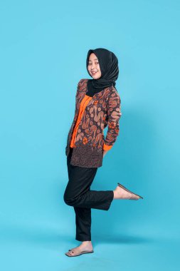 Neşeli genç Asyalı Müslüman kadın bir bacağını kaldırırken mutlu bir şekilde gülümsüyor, geleneksel batik kıyafetler ve başörtüsü giyiyor, parlak mavi bir arka plana karşı güvenle duruyor..