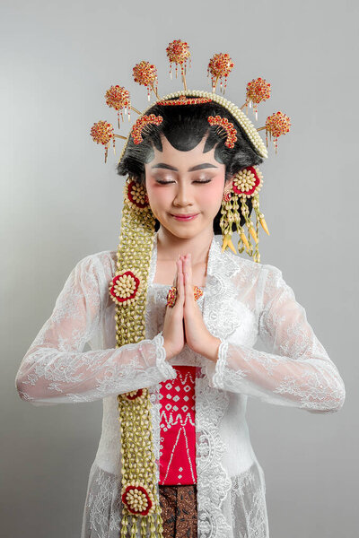 An Indonesian bride in Solo Putri attire stands gracefully with a soft smile, hands in a respectful greeting. She wears a golden headdress, jasmine garland, and a white lace kebaya.