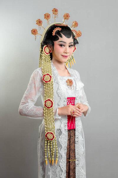 Indonesian bride in Solo Putri attire, wearing an ornate golden headdress, jasmine garlands, and a white lace kebaya. She poses elegantly with a soft, charming smile.