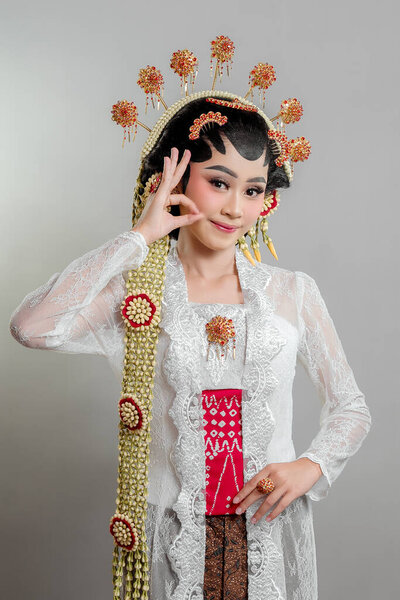 Indonesian bride in Solo Putri attire, wearing a lace kebaya, intricate headdress, and jasmine garlands. She smiles gracefully while making an "OK" hand gesture.