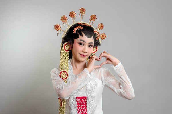 Indonesian bride in Solo Putri attire, wearing a lace kebaya, intricate headdress, and jasmine garlands. She smiles sweetly while forming a heart shape with her hands.