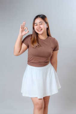 İki kere tamam el hareketi yaparken neşeli, genç bir kadın ışıl ışıl gülümsüyor. Neşeli yüz ifadesi, reklam kullanımı için güven, anlaşma ve pozitif iletişim sağlıyor..