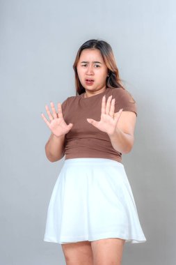 Asyalı genç bir kadın kendini savunmak için ellerini kaldırarak şok ve korku ifade ediyor. Güçlü yüz tepkisi, geniş gözler ve açık ağız, gri arka planda izole edilmiş, duygusal ifade kavramı.