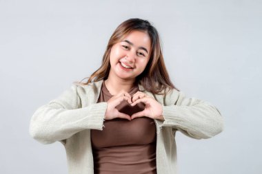 Samimi bir süveter giyen gülümseyen genç bir kadın elleriyle bir kalp yapar. Tarafsız bir arkaplana karşı sevgi, şefkat ve şefkat ifade eder..
