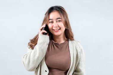 Rahat bir süveter giyen genç bir kadın cep telefonuyla konuşurken gülümsüyor. Tarafsız bir arkaplanda iletişim ve bağlantı kuruyor.