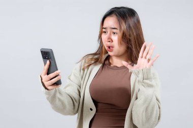 Genç bir kadın akıllı telefonuna bakarken hayal kırıklığını gösterir. Tarafsız bir arkaplanda güçlü bir duygusal tepki gösterir..