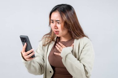 Samimi bir süveter giyen genç bir kadın cep telefonunda gördüğü bir şeye şaşırır ve şaşırır. Eli göğsünde, tarafsız bir arkaplanda..