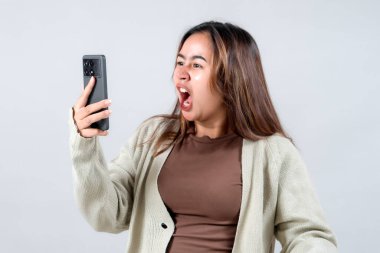 Genç bir kadın akıllı telefonuna bakarken hayal kırıklığını gösterir. Tarafsız bir arkaplanda güçlü bir duygusal tepki gösterir..