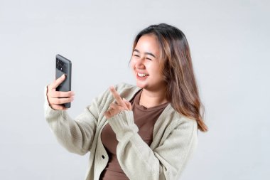 Genç bir kadın gülümser ve akıllı telefonuna işaret eder. Tarafsız bir arkaplanda cihazıyla uğraşırken neşe ve heyecan ifade eder..