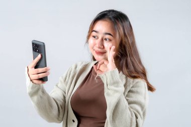 Genç bir kadın gülümsüyor ve akıllı telefonuna el sallıyor. Tarafsız bir arkaplanda görüntülü konuşma yaparken arkadaşlığını ve sıcaklığını ifade ediyor.