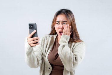 Genç bir kadın akıllı telefonuna bakarken şok ve güvensizlik gösteriyor, tarafsız bir arkaplanda ağzını şaşkınlıkla kapatıyor..