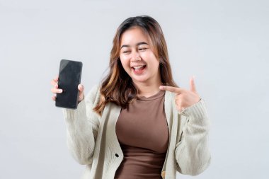 Genç bir kadın gülümsüyor ve akıllı telefonuna işaret ediyor. Cihazı tarafsız bir arkaplanda tutarken heyecan ve coşku gösteriyor..