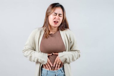 Genç bir kadın, rahat bir süveter giyerek karnını tutarken rahatsız edici bir şekilde somurtuyor. Yüzündeki ifade, acı ya da endişeye işaret ediyor, anlaşılmaz bir ıstırap anını yakalıyor..