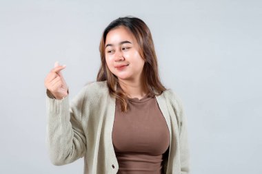 Genç bir kadın, genellikle sevgi ve şefkat ile ilişkilendirilen neşeli bir el hareketi yaparken ışıl ışıl gülümser. Samimi bir süveter giyiyor. Tarafsız bir zemine karşı neşeli ve dostane bir hava yayıyor.