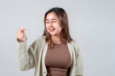 Genç bir kadın, genellikle sevgi ve şefkat ile ilişkilendirilen neşeli bir el hareketi yaparken ışıl ışıl gülümser. Samimi bir süveter giyiyor. Tarafsız bir zemine karşı neşeli ve dostane bir hava yayıyor.