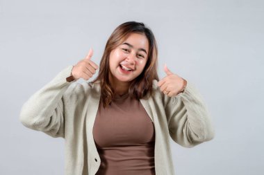 Genç bir kadın sevinçle ışıldıyor, neşeli bir jestle iki parmağını kaldırıyor. Samimi bir süveter giymişti, parlak gülüşü ve pozitif enerjisiyle sade gri bir arka planda parlıyordu, mutluluk saçıyordu.