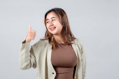 Genç bir kadın sevinçle ışıldıyor, neşeli bir jestle başparmağını kaldırıyor. Samimi bir süveter giymişti, parlak gülüşü ve pozitif enerjisiyle sade gri bir arka planda parlıyordu, mutluluk saçıyordu.