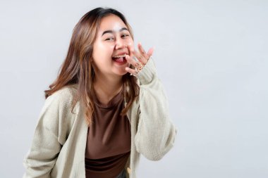 Genç bir kadın yürekten güler, eli şakacı bir hareketle ağzını sıvazlar. Kazak giyiyor, neşeli bir ifade takınıyor ve gülümsüyor. Tarafsız bir arkaplanda mutluluk ve eğlence yayıyor..