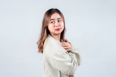 Sıcak bir ifadeyle gülümseyen genç bir kadın, rahat bir süveter giyiyor, ellerini kalbine yakın tutuyor, nötr bir arkaplana karşı kibarlık ve pozitiflik yayıyor..