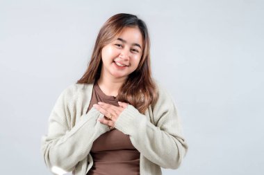 Sıcak bir ifadeyle gülümseyen genç bir kadın, rahat bir süveter giyiyor, ellerini kalbine yakın tutuyor, nötr bir arkaplana karşı kibarlık ve pozitiflik yayıyor..