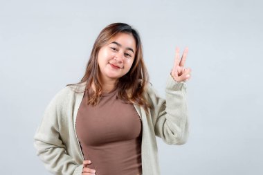 Rahat bir süveter giyen neşeli genç bir kadın, tarafsız bir arkaplana karşı olumlu ve neşeli bir ruh yayarak barış işareti yapar..