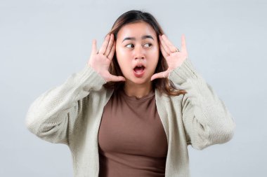 Rahat bir süveter giyen genç bir kadın, elleri çerçeveli ve ağzı açık, tarafsız bir arka plana karşı şaşkınlık ve şok ifade eder..