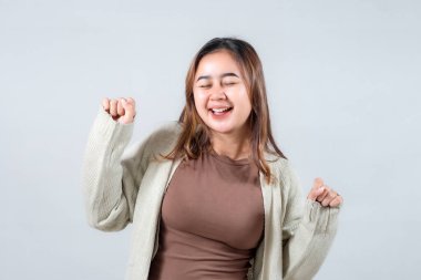 Samimi bir süveter giyen genç bir kadın neşeyle dans eder, gözleri kapalı ve yüzünde parlak bir gülümseme, tarafsız bir arka plana karşı mutluluk ve tasasız ruhu ifade eder..