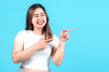 Beyaz tişörtlü gülümseyen genç bir kadın iki eliyle şakacı bir şekilde işaret ediyor, parlak mavi bir arka plana karşı neşeli bir tavır sergiliyor, içeriği meşgul etmek için mükemmel