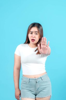 Beyaz tişörtlü, şaşırmış genç bir kadın elini kaldırıyor. Durdurucu bir jest yapıyor, parlak mavi bir arka plana karşı şok ya da hoşnutsuzluğu ifade ediyor, güçlü duygular taşıyor.