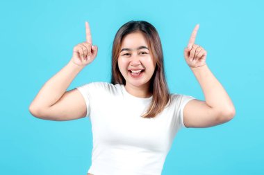 Beyaz tişörtlü neşeli genç bir kadın, geniş bir gülümseme ve her iki elini kaldırıyor işaret parmakları yukarı, parlak mavi bir arka plan karşı kurulmuş, pozitif yayılıyor.