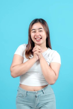 Heyecanlı genç bir kadın elini sıkar, coşkulu mavi bir arka plana sevinçle ışıldar, mutluluk ve beklenti ifade eder.