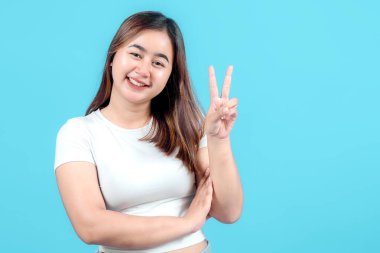 Beyaz ekin bluzlu mutlu genç bir kadın, parlak mavi bir arka plana gülümseyen, neşe ve pozitiflik saçan, eliyle barış işareti yapıyor..