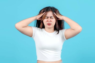 Beyaz bir bluz giymiş genç bir kadın şok ve acıyla tepki veriyor, kafasını mavi bir arka plana yaslayarak, hayal kırıklığını veya paniği ifade ediyor.