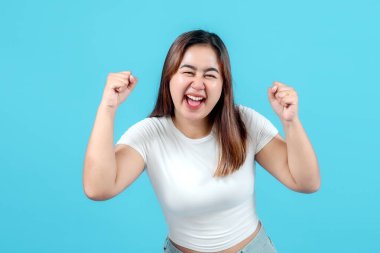 Neşeli genç bir kadın mavi bir arka plana yumruk sallayarak mutluluğu ve heyecanı ifade ederek kutluyor. Başarı, pozitiflik ve başarı temaları için mükemmel..