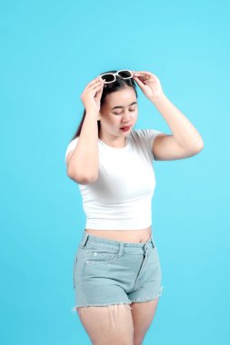 Beyaz bir bluz ve kot pantolon giyen genç bir kadın neşeli bir şekilde güneş gözlüklerini parlak bir mavi arka plana göre ayarlıyor. Eğlenceli ve kaygısız bir yaz havası yayıyor..