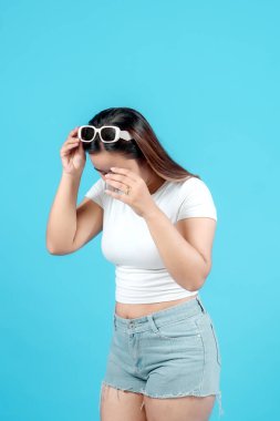 Beyaz bir bluz ve kot pantolon giyen genç bir kadın neşeli bir şekilde güneş gözlüklerini parlak bir mavi arka plana göre ayarlıyor. Eğlenceli ve kaygısız bir yaz havası yayıyor..