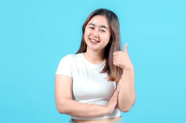 Beyaz tişörtlü gülümseyen genç bir kadın olumlu bir jest yapıyor, parlak mavi bir arka plana karşı pozitif tutumunu sergiliyor, kendine güven ve mutluluk yayıyor..