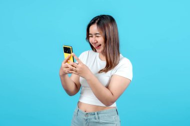 Beyaz tişörtlü genç bir kadın, neşeli bir şekilde sarı akıllı telefonuna bakıyor, açık mavi bir arka plana gülümsüyor, mutluluk ve nişan anını yakalıyor.