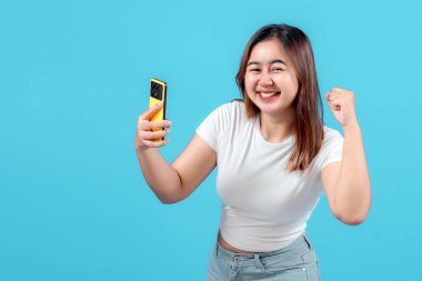 Neşeli genç bir kadın, canlı mavi bir arka plana karşı heyecan ve mutluluk sergileyerek akıllı telefonuyla kutlama yapıyor..