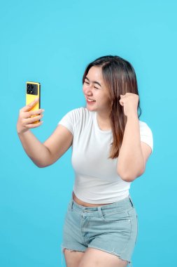 Beyaz tişörtlü heyecanlı genç kadın, sarı akıllı telefonuna bakarak, parlak mavi bir arka plana karşı neşe ve zafer ifade ederek kutlama yapıyor.