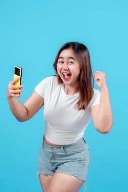 Neşeli genç bir kadın, canlı mavi bir arka plana karşı heyecan ve mutluluk sergileyerek akıllı telefonuyla kutlama yapıyor..