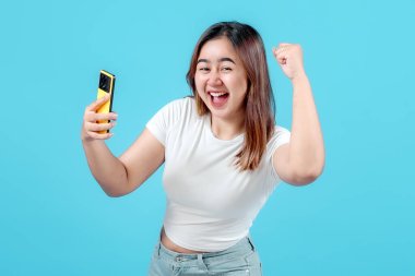 Neşeli genç bir kadın, canlı mavi bir arka plana karşı heyecan ve mutluluk sergileyerek akıllı telefonuyla kutlama yapıyor..