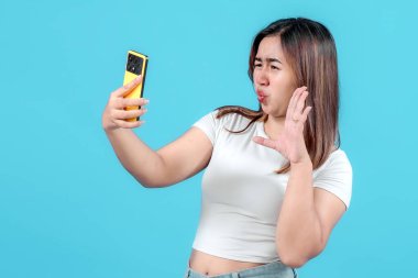 Şaşırmış genç bir kadın, akıllı telefonuna bakarken şokla tepki verir. Parlak mavi bir arka plana karşı kuşku ve heyecanı ifade eder..