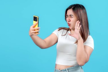 Şaşırmış genç bir kadın, akıllı telefonuna bakarken şokla tepki verir. Parlak mavi bir arka plana karşı kuşku ve heyecanı ifade eder..