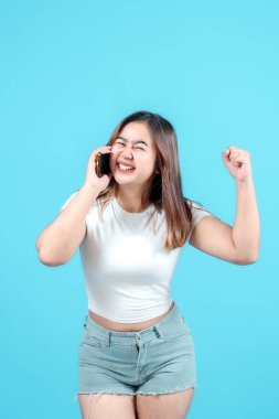 Beyaz tişörtlü heyecanlı genç kadın, parlak mavi bir arka plana karşı neşe ve başarısını ifade ederek akıllı telefonuyla konuşurken iyi haberleri kutluyor.