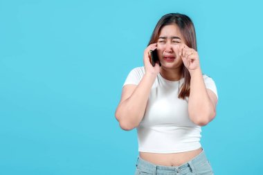 Sıkıntılı genç bir kadın, parlak mavi bir arka plana karşı üzüntü ve hayal kırıklığını ifade ederek, akıllı telefonuyla konuşurken ağlar..