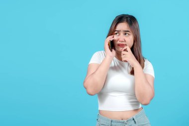 Endişeli genç bir kadın, parlak mavi bir arka planda parmağını ısırırken endişeli bir yüz ifadesini göstererek akıllı telefonuyla konuşur..