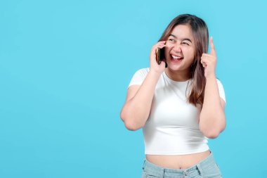 Neşeli genç bir kadın akıllı telefonuyla konuşuyor, parlak mavi bir arkaplanı işaret ederken mutlu bir yüz ifadesi sergiliyor..